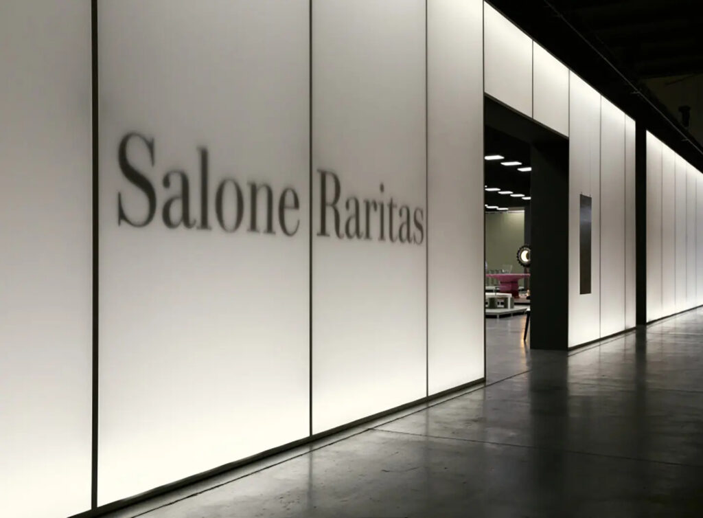 Entrada iluminada do Salone Raritas com branding tipográfico em parede branca moderna, acesso a área de exposição de peças raras e colecionáveis de design.