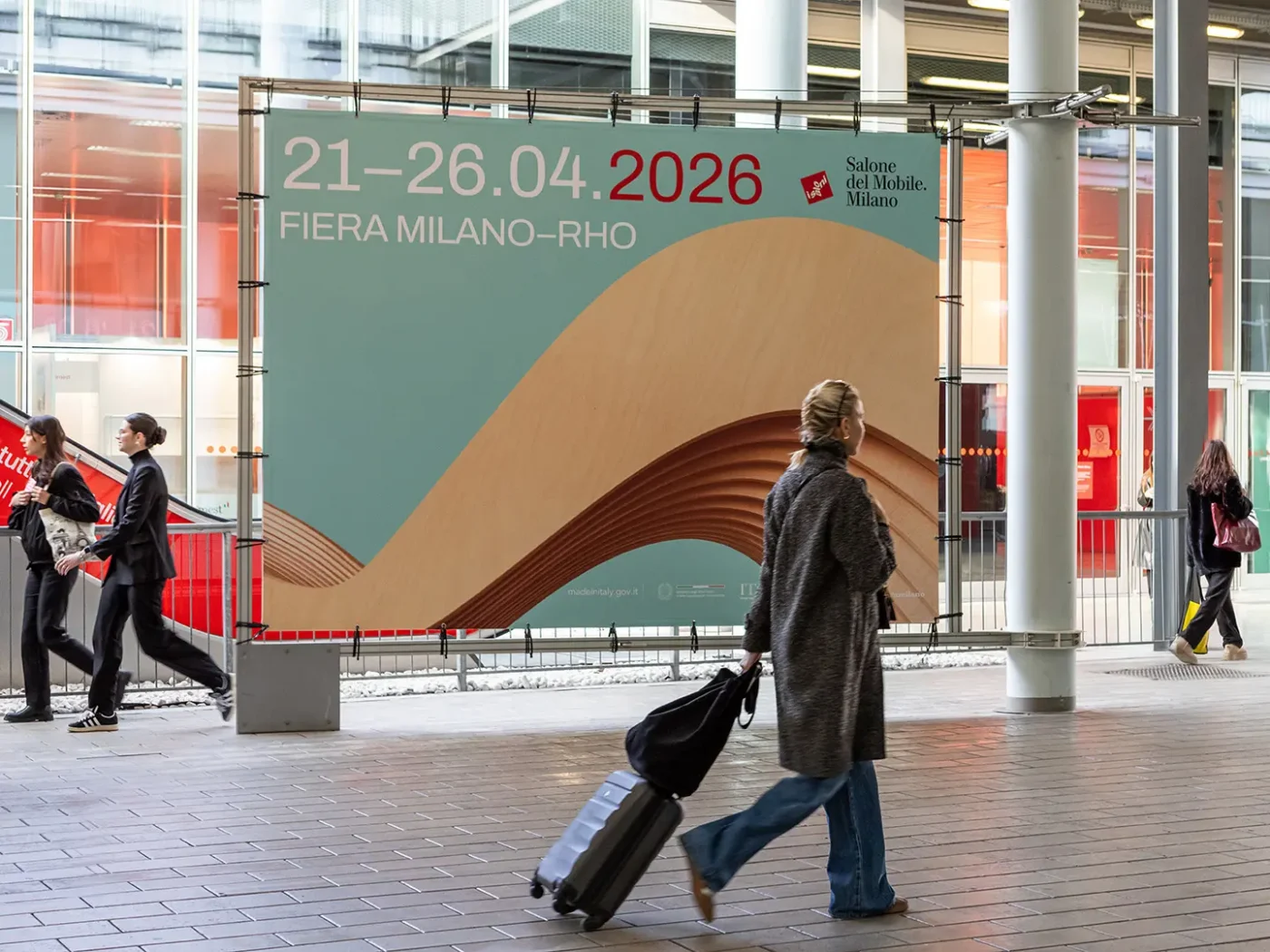 Convite visual para o Salone del Mobile 2026 com datas e localização, capturando momento de chegada de participante com mala, simbolizando a experiência internacional.