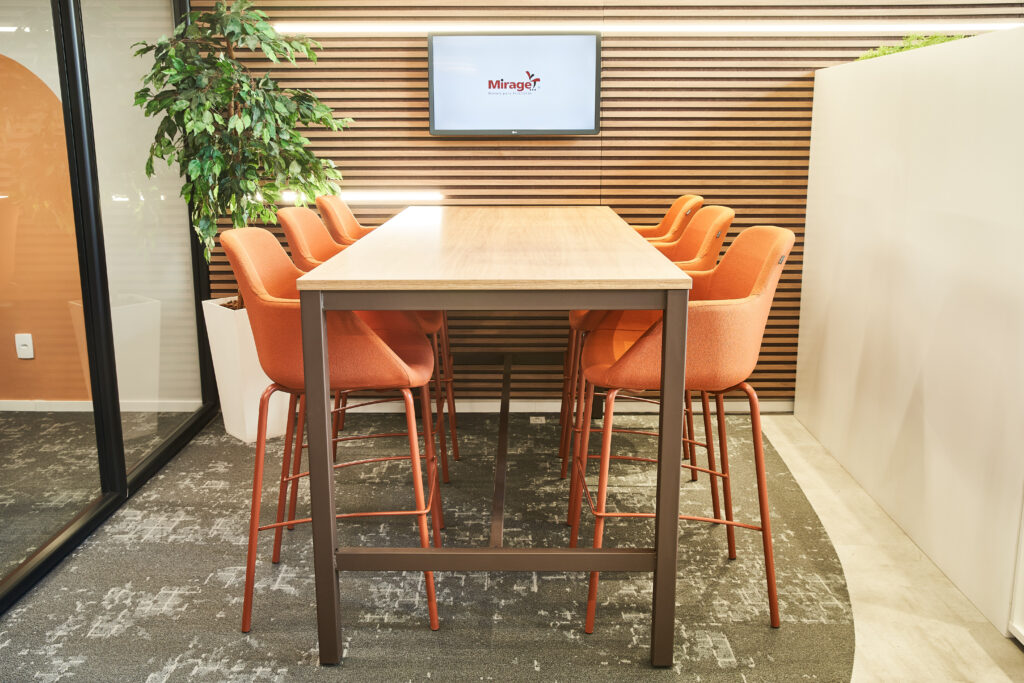 Ambiente corporativo planejado pela Mirage Móveis com mesa alta, cadeiras laranja, painel ripado e soluções de mobiliário corporativo.